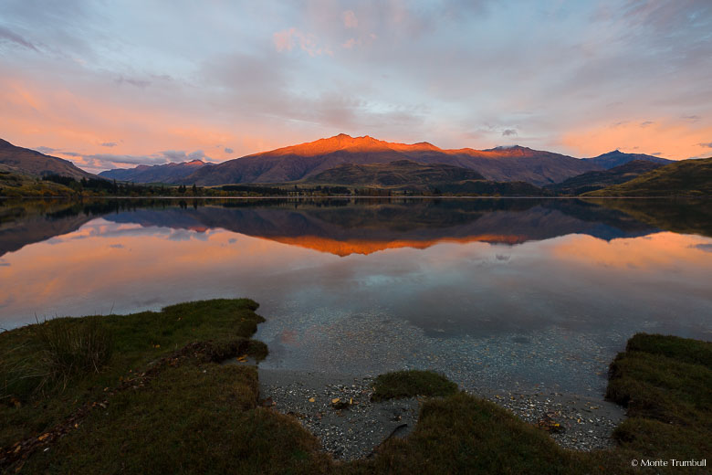 The rising sun paints the clouds pink and illuminates the top of Mount Hyde and is reflected in the still water of Glendhu Bay on Lake Wanaka on the South Island of New Zealand.