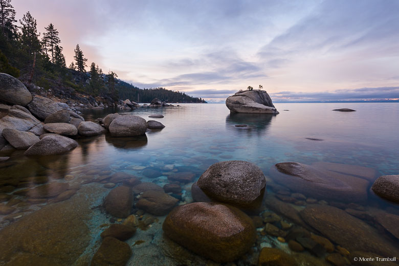 The pink skies of dawn reflect in the calm water around Bonsai Rock at Lake Tahoe in Nevada.