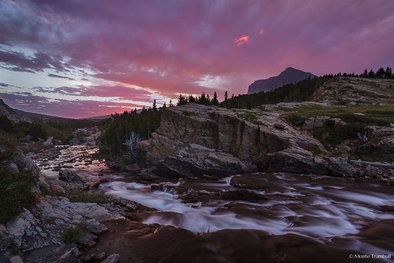 Swiftcurrent Creek rushes down Swiftcurrent Falls and flows down a valley towards the rising sun and a sky full of pink clouds in Glacier National Park, Montana.