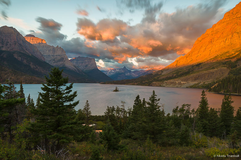 The rising sun streams across the tops of the mountains surrounding Saint Mary Lake painting the clouds and the mountainsides orange on a windy morning in Glacier National Park, Montana.